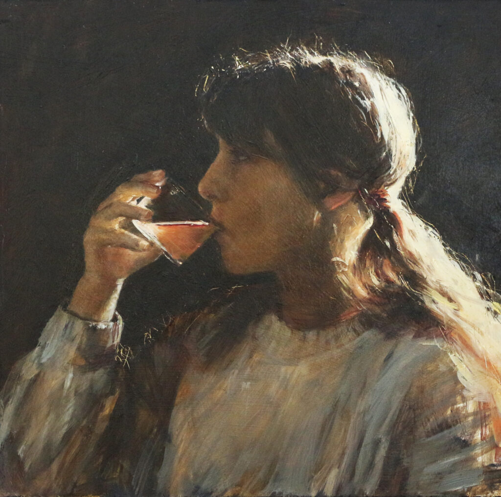 Portrait of a young girl by Ros Koch. She is sitting with her back to the light, darkness behind her, sipping an amber-colured liquid from a tumbler glass. She is wearing a grey jumper, with her hair tied back in a pony tail and the crown of her head is backlit from behind.
